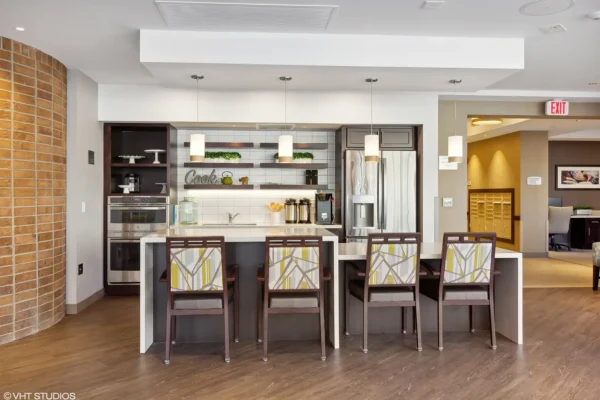 Modern kitchen area with a white island, four patterned chairs, pendant lighting, open shelves with decor, and built-in appliances.