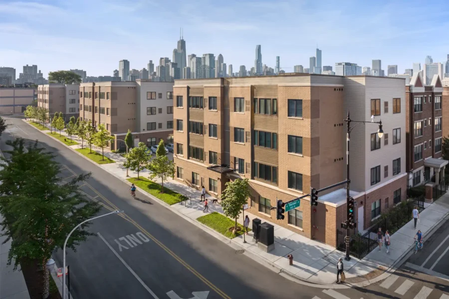 A modern apartment complex on a street corner with pedestrians, trees lining the sidewalk, and a city skyline in the background.