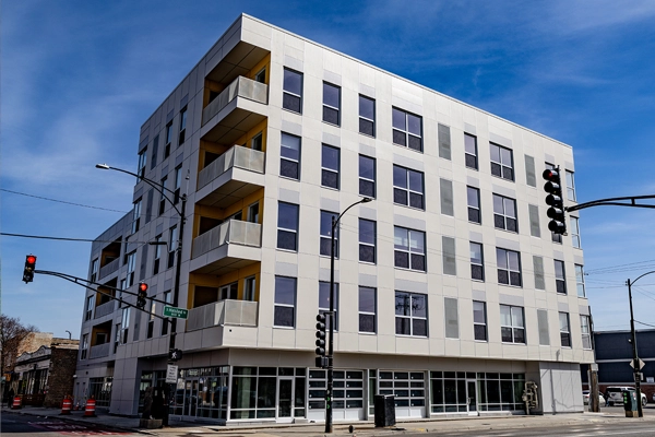 A modern five-story apartment building with large windows and balconies, located at a street corner with traffic lights and clear skies.