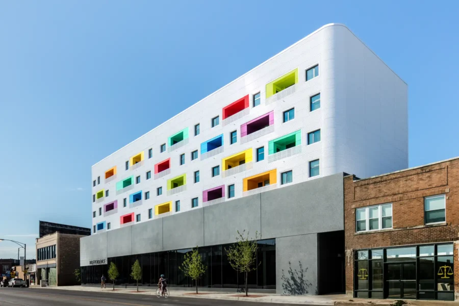 A modern white building with rectangular, colorful window frames sits above a gray ground floor on a city street with a few pedestrians and young trees.