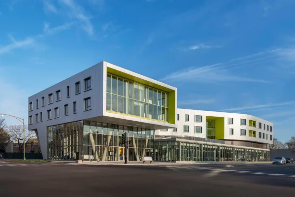 Modern building with large glass windows and white geometric exterior, featuring green accents, situated at a street corner under a clear blue sky.