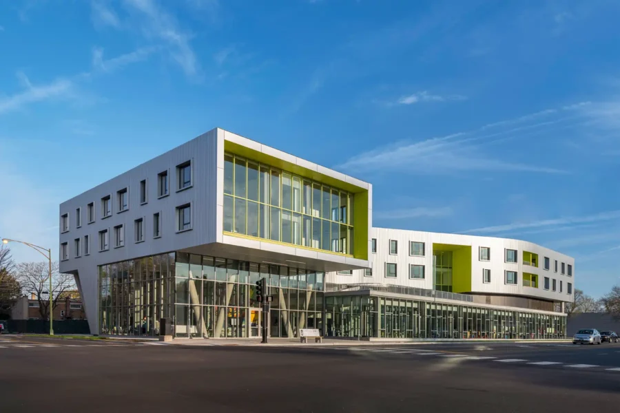 Modern building with large glass windows and white geometric exterior, featuring green accents, situated at a street corner under a clear blue sky.