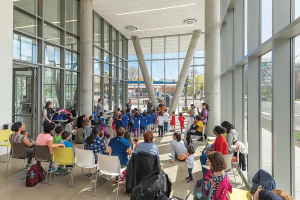 A group of adults and children are gathered in a spacious, sunlit room with large windows, seated and standing, watching a group activity in the center.