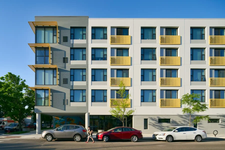 Four-story modern apartment building with yellow balcony railings, large windows, and ground-floor parking. Three cars are parked in front, and two people walk on the sidewalk.