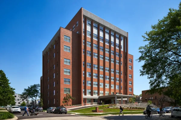 A modern, multi-story red brick office building with large windows stands on a landscaped corner lot under a clear blue sky. Pedestrians and parked cars are visible nearby.