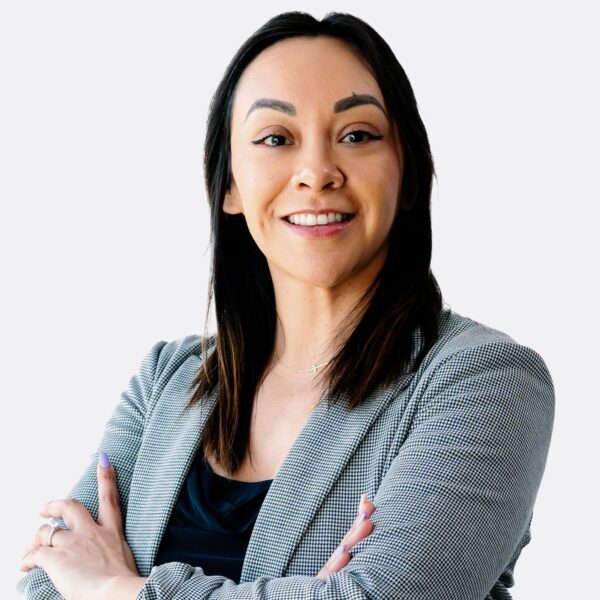 Woman with long dark hair wearing a gray blazer and black top, standing with arms crossed and smiling at the camera against a plain light background.