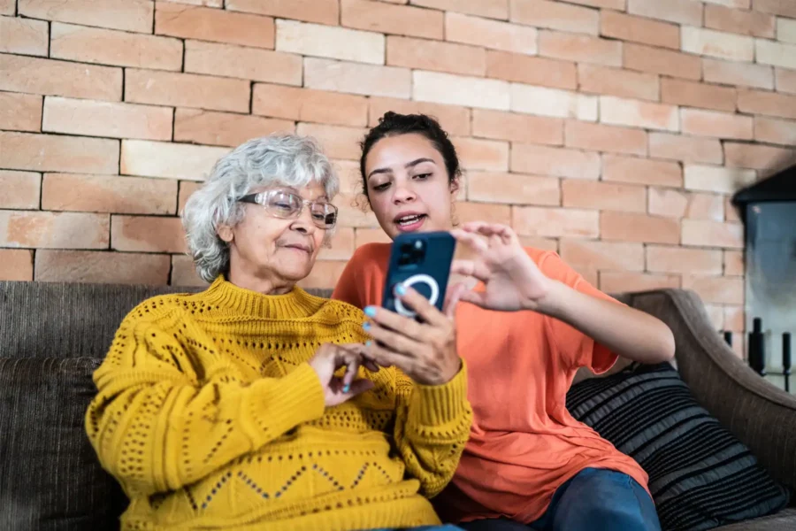 An older woman and a younger woman sit on a couch; the younger woman helps the older woman use a smartphone.