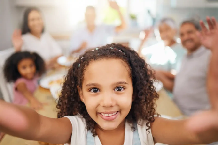 Young girl smiling at the camera with her arms outstretched, while four adults and another child sit at a table in the background, all smiling and waving.