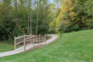 A wooden footbridge crosses over a small gravel path, surrounded by green grass, trees, and dense foliage in a park-like setting.
