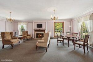 Formal living room with armchairs, dining table, fireplace, TV, chandeliers, large windows, and light pink walls.