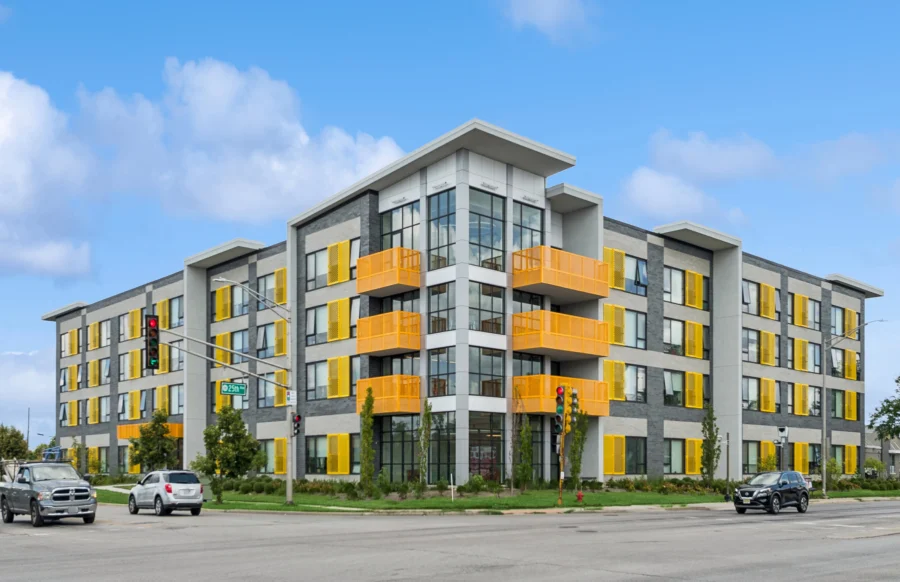 Four-story modern apartment building with gray and white exterior, large windows, and prominent yellow balcony railings on a street corner with cars and traffic lights.