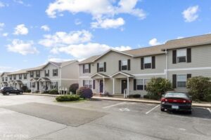 Row of two-story beige townhouses with small porches, shrubs, and a parking lot with cars and a handicap space under a partly cloudy sky.