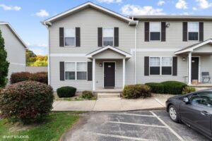 A two-story beige townhouse with black shutters, a small covered entryway, and landscaped bushes in front, next to a parking lot with a car parked nearby.