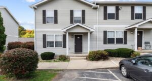 A two-story townhouse with gray siding, black shutters, and a black door; a small concrete porch, shrubs, and a parked car are visible in front.