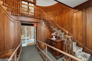 Wood-paneled foyer with curved staircase, wooden railing, floral-upholstered bench, chest of drawers with vase of flowers, and a ramp leading to a doorway.