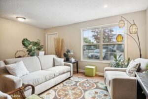 Bright living room with beige walls, two sofas, green plants, a patterned rug, a window letting in daylight, and a modern floor lamp with three shades.