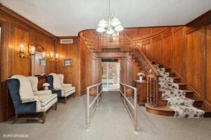 Wood-paneled foyer with carpeted curved staircase, chandelier, accessibility ramp, and armchairs along the wall.