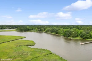 A calm river winds through green marshland and forested areas under a partly cloudy sky, with docks along the water’s edge.