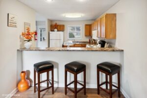 A kitchen with a breakfast bar, three black stools, wooden cabinets, white appliances, and decorative items including a vase with orange flowers.
