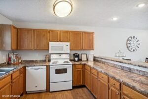 Kitchen with wood cabinets, white appliances including stove, microwave, and dishwasher, marble-look countertops, and a wall clock in the background.