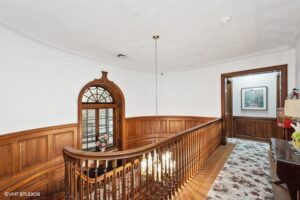 Second-floor hallway with wooden paneling, curved banister, hanging chandelier, floral carpet, and framed wall art in a traditional-style home.