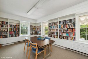 A bright library room with built-in bookshelves on three walls, four chairs around a rectangular table, and two windows letting in natural light.
