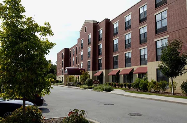 A four-story brick building with large windows and red awnings, featuring a covered entrance and landscaping along a paved driveway.