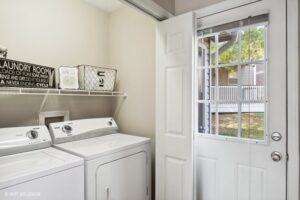 A laundry room with a washer and dryer, wire shelf holding baskets, and an open door leading to a yard outside.