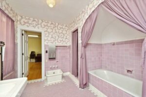 Bathroom with pink tile walls, matching pink fixtures, a bathtub with a pink curtain, and floral wallpaper. Door opens to an adjacent room with wooden floor and chairs.