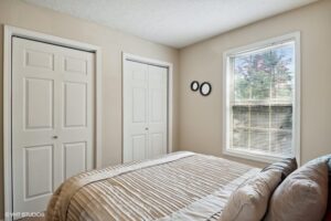 A small bedroom with a neatly made bed, two closets with white doors, beige walls, and a window with blinds letting in natural light.