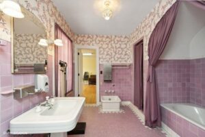 Bathroom with pink floral wallpaper, pink tiles, a white sink, bidet, bathtub with curtain, and white fixtures. Doorway leads to a room with wood flooring.