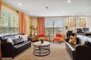 Modern living room with two black sofas, two orange chairs, a round coffee table, large windows with drapes, abstract wall art, and a desk with a computer in the background.