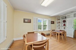 A dining room with two wooden tables and chairs, large windows with white shutters, built-in shelves displaying decor, and light yellow walls.
