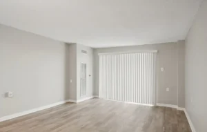 Empty room with light gray walls, wood-style flooring, and vertical blinds covering a large window or sliding glass door.