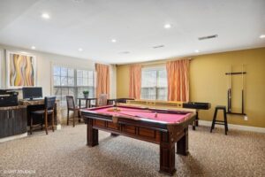 A room with a pool table, computer desk, small table with chairs, wall-mounted cues, and large windows with orange curtains.