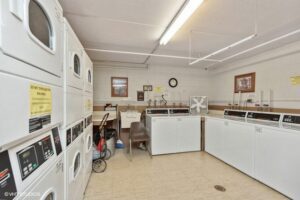 A laundry room with multiple washers and dryers, a sink, a chair, a cart, and a box fan against the wall, under fluorescent lighting.