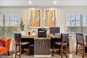 A small office area with two chairs, a granite-top desk, a computer, a printer, decorative plants, and two abstract art pieces on the wall between two large windows.