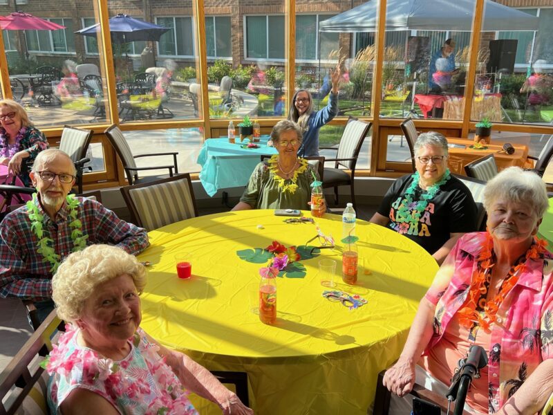 A group of seniors sit around a yellow table decorated with leis, drinks, and flowers at a sunlit indoor gathering. A woman in the back waves at the camera.