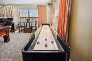 A shuffleboard table is positioned in a carpeted room with large windows, orange patterned curtains, and a table with chairs in the background.