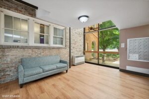 A small lobby area with a blue sofa, brick walls, wood floor, large window, mailboxes, and a view of trees and a brick arch outside.
