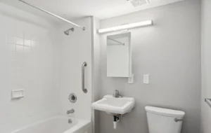 A simple bathroom with a white bathtub and shower, a grab bar, a wall-mounted sink, a mirror, a toilet, and overhead lighting.