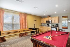 Game room with a red pool table, shuffleboard, foosball, kitchenette with microwave and mini fridge, large window with orange curtains, and neutral walls.