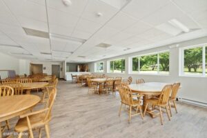 A spacious dining area with round wooden tables and chairs, large windows on one side, and light wood flooring.