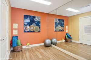 A small exercise room with wood flooring, orange accent wall, a large wall mirror, two yoga balls, rolled yoga mats, and a colorful abstract artwork.
