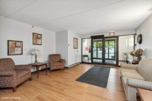 Lobby area with two armchairs, a sofa, side tables, framed art on white walls, a lamp, floral arrangement, and glass entrance doors with a view outside.
