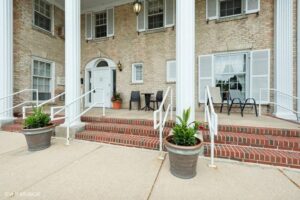 Brick building entrance with white columns, steps, two large potted plants, and outdoor seating with black chairs and a table near shuttered windows.