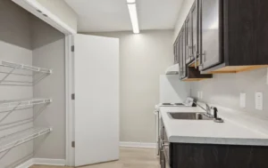 Narrow kitchen with dark cabinets, white countertop, stainless steel sink, electric stove, and an open door revealing empty wire shelving.