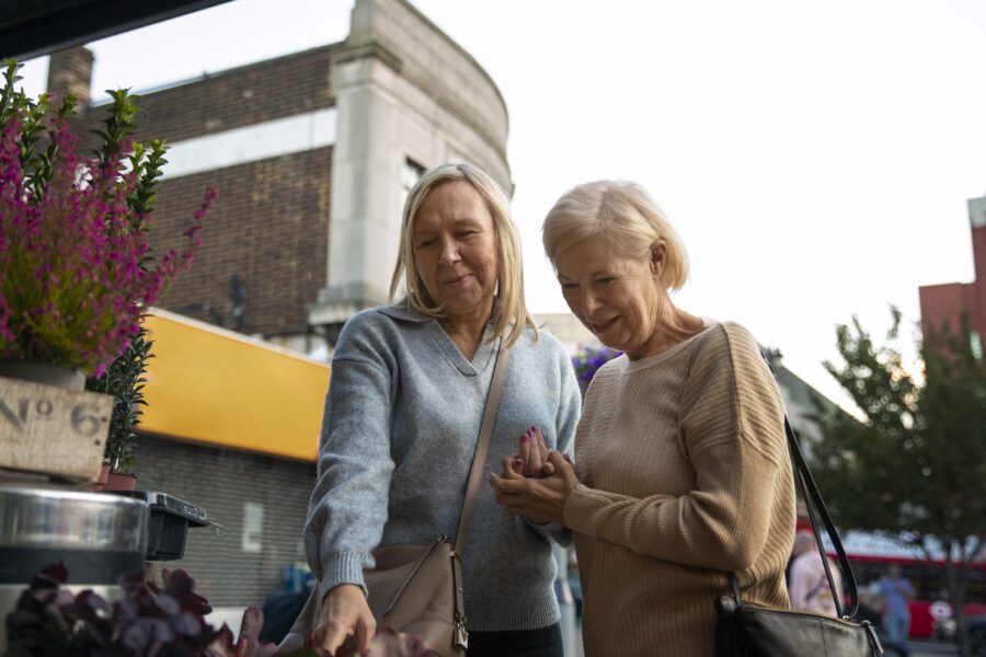 Two women stand close together at an outdoor market, looking at plants on display. They are both casually dressed and appear engaged in conversation.
