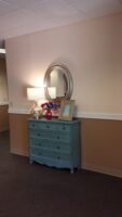 A light blue dresser with a lamp, mirror, framed photo, and small patriotic decorations stands against a beige wall in a hallway.