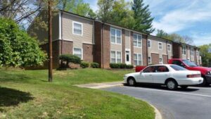 A two-story brick and siding apartment building with parked cars in front, bordered by grass and trees under a partly cloudy sky.